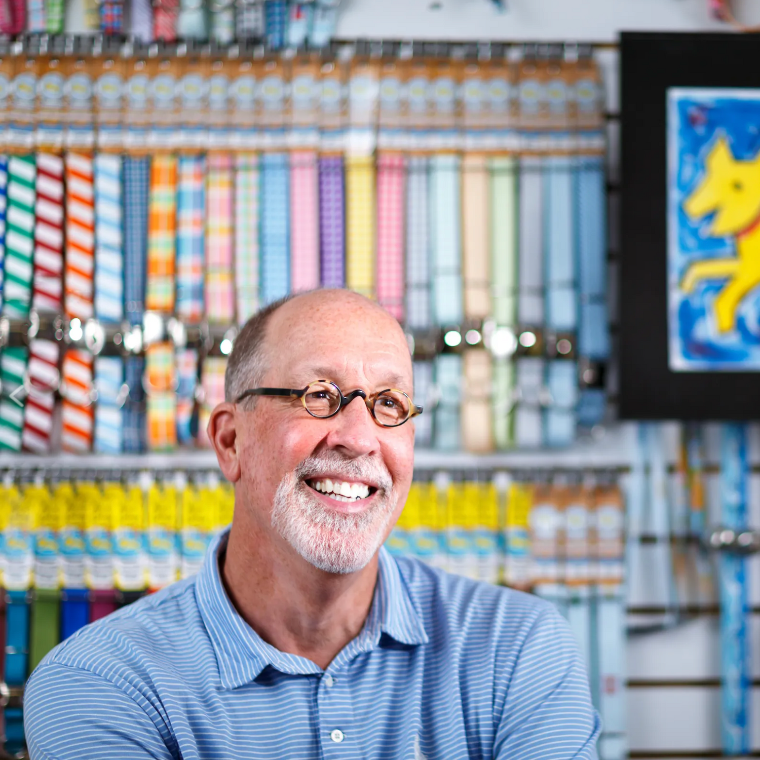 Man smiling in a store with colorful products and a painting of a dog on a screen.