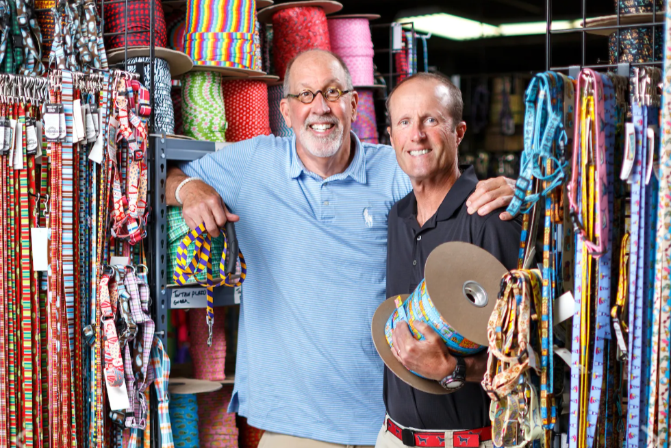 Two men standing in a store with colorful ribbon spools and belts around them.